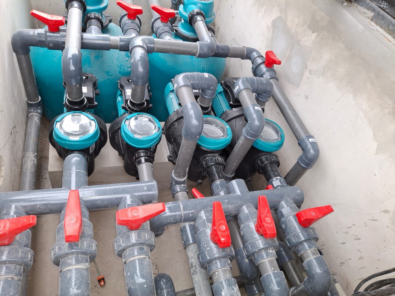 Sistema de tuberías y bombeo con manijas de válvulas rojas, dentro de un recinto de hormigón.