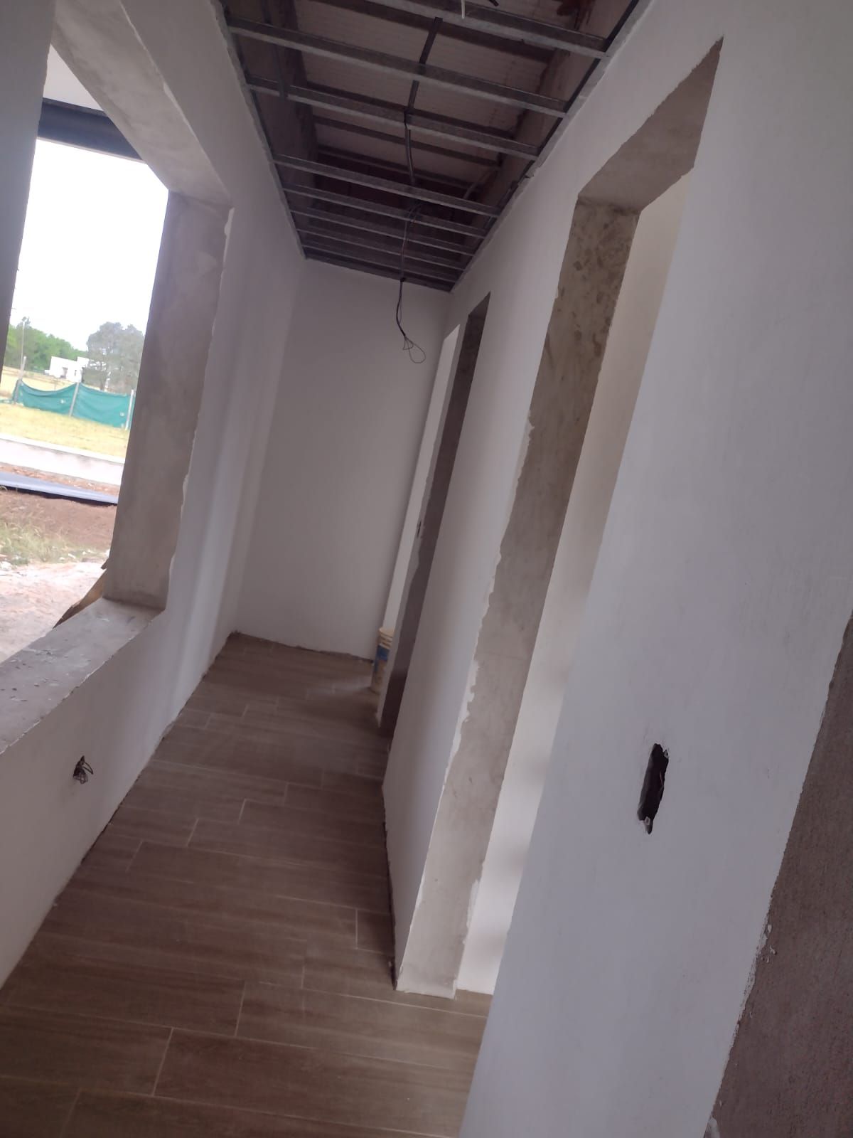 Pasillo interior en construcción con techo sin terminar, paredes blancas, suelo con aspecto de madera y puertas.