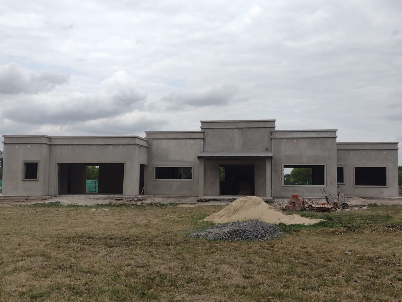 Casa en construcción; exterior de hormigón con ventanas y puertas sin terminar, situada en un campo.