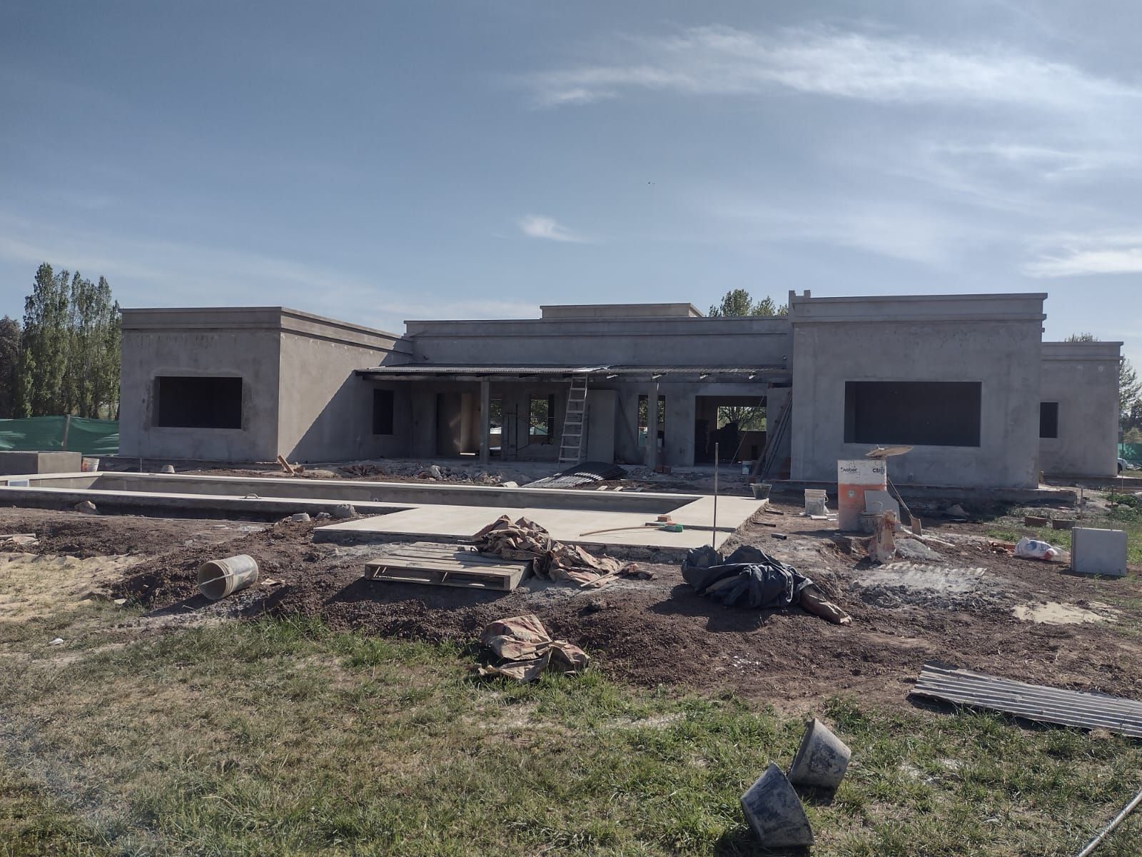 Casa en construcción en un terreno cubierto de hierba, estructura de hormigón sin terminar, cielo azul.