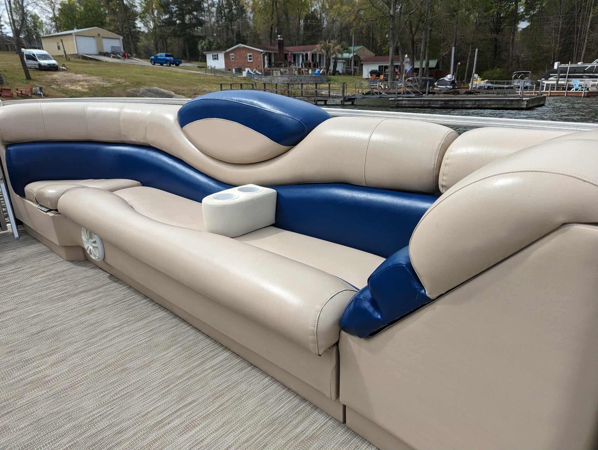 Beige and blue pontoon boat seating with a built-in cupholder, by a body of water.