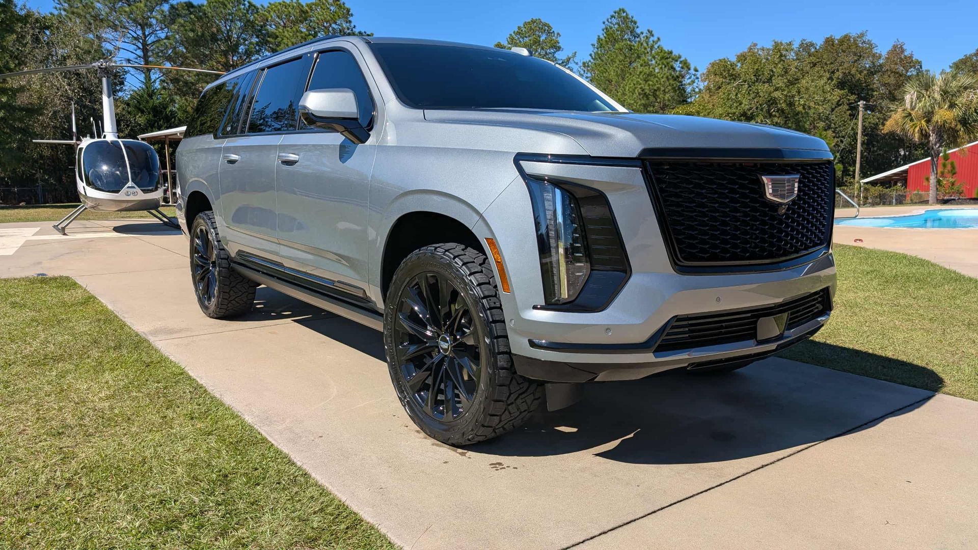 Silver Cadillac SUV on a concrete driveway with a helicopter in the background. Black wheels, tinted windows, sunny day.