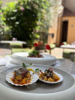 Due capesante servite nel guscio su un piatto bianco, con sullo sfondo un giardino esterno sfocato.