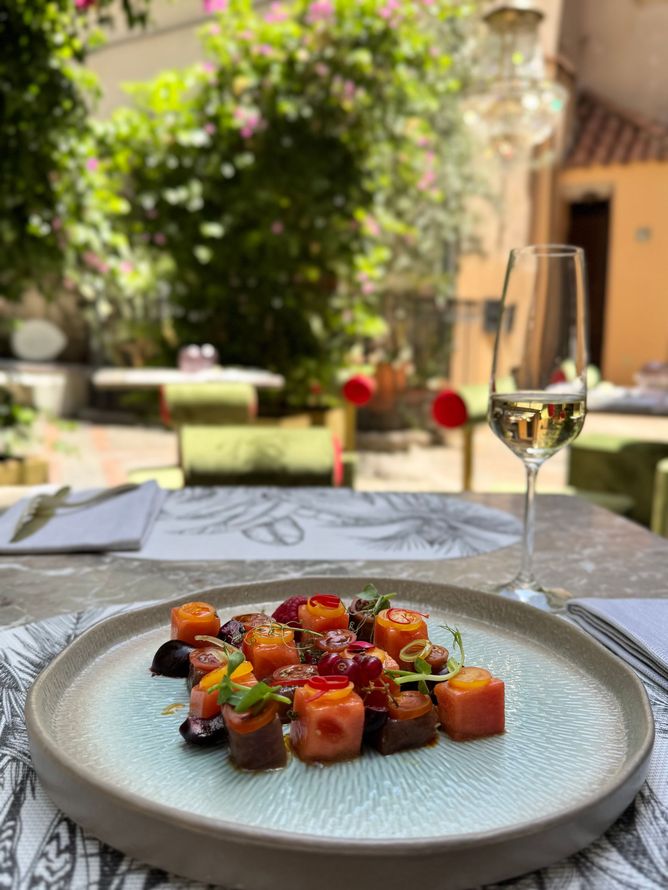 Un piatto gourmet a base di salmone a cubetti e insalata di barbabietole, servito su un piatto dalla consistenza particolare, accanto a un bicchiere di vino, in un cortile soleggiato.