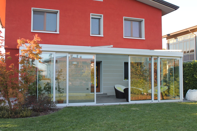 Veranda con vetrate annessa a una casa rossa, con porte scorrevoli in vetro che si aprono su un prato.