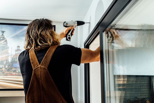 Una persona con una tuta marrone fora un muro vicino al telaio di una finestra nera; ambiente interno.