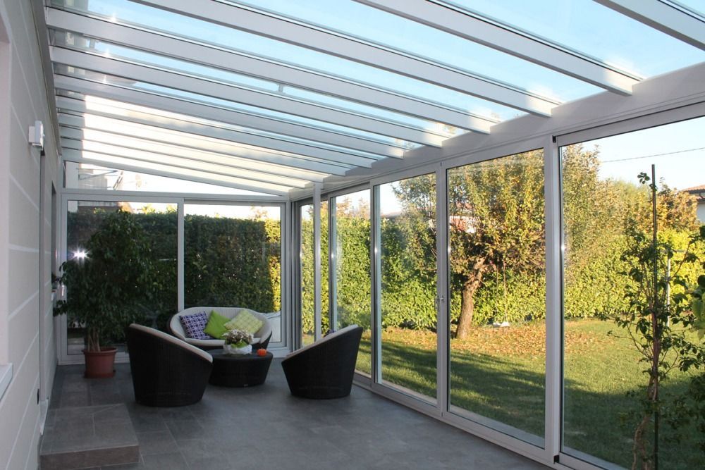 Veranda con struttura bianca, pareti in vetro e vista sul verde del giardino. Arredata con sedie e divano.