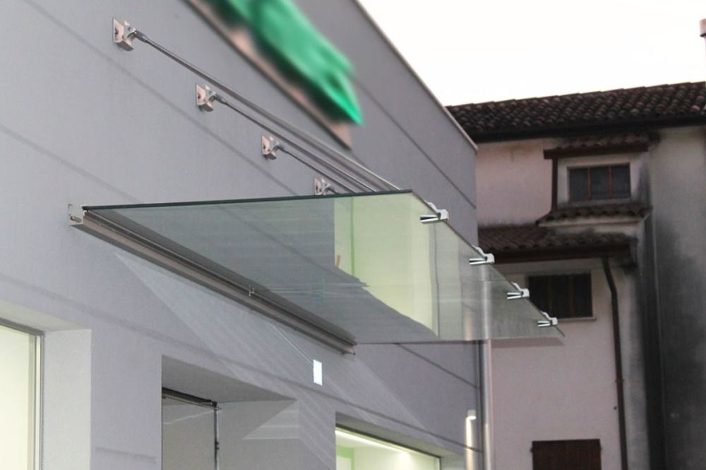 Tenda in vetro fissata alla facciata di un edificio, con supporti e cavi metallici, che protegge l'ingresso.