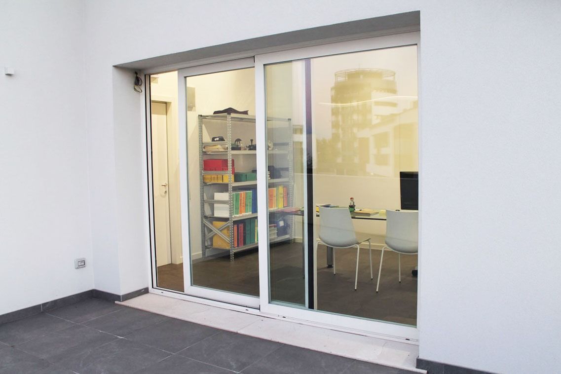 Porta scorrevole in vetro bianco che conduce a una stanza con scaffali, scrivania e sedie; la porta è aperta.