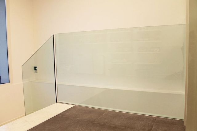 Parete doccia in vetro con sezione inclinata, adiacente a una parete, pavimento in piastrelle beige.