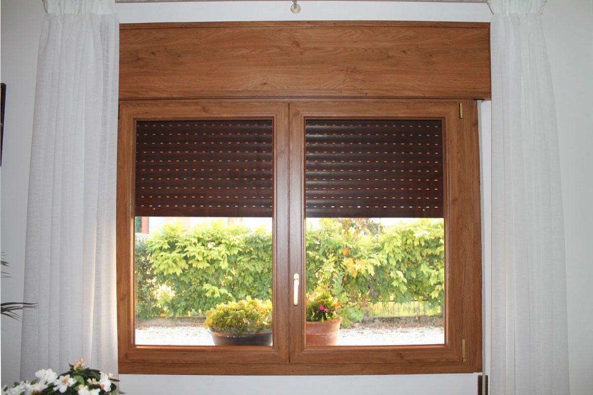 Finestra marrone con persiane chiuse e tende bianche trasparenti. Foglie verdi visibili all'esterno.