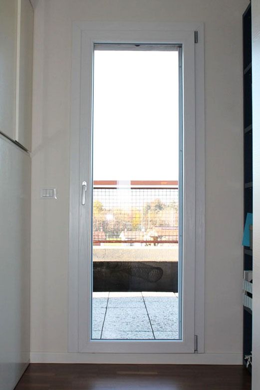 Porta con cornice bianca che si apre su un patio. Pavimento marrone all'interno. Luce solare intensa.