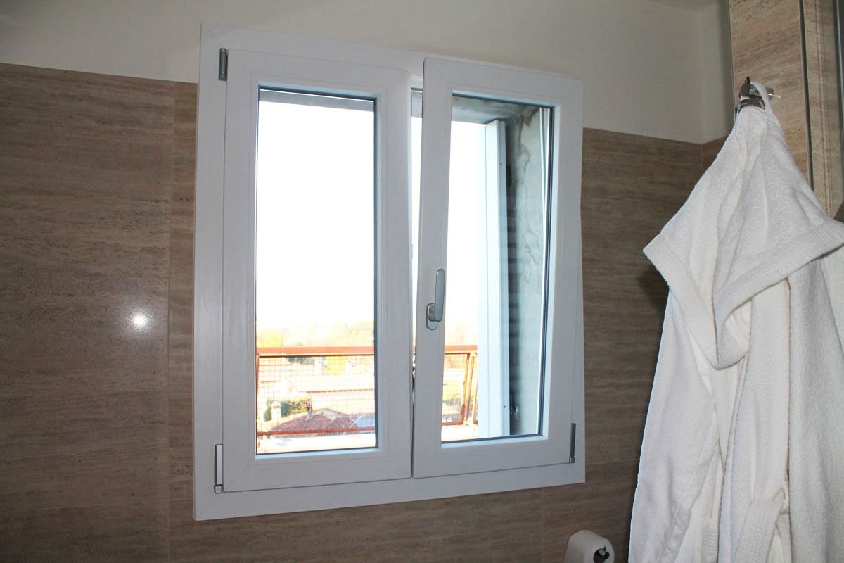 Finestra bianca in un bagno con accappatoio bianco appeso, pareti piastrellate beige.