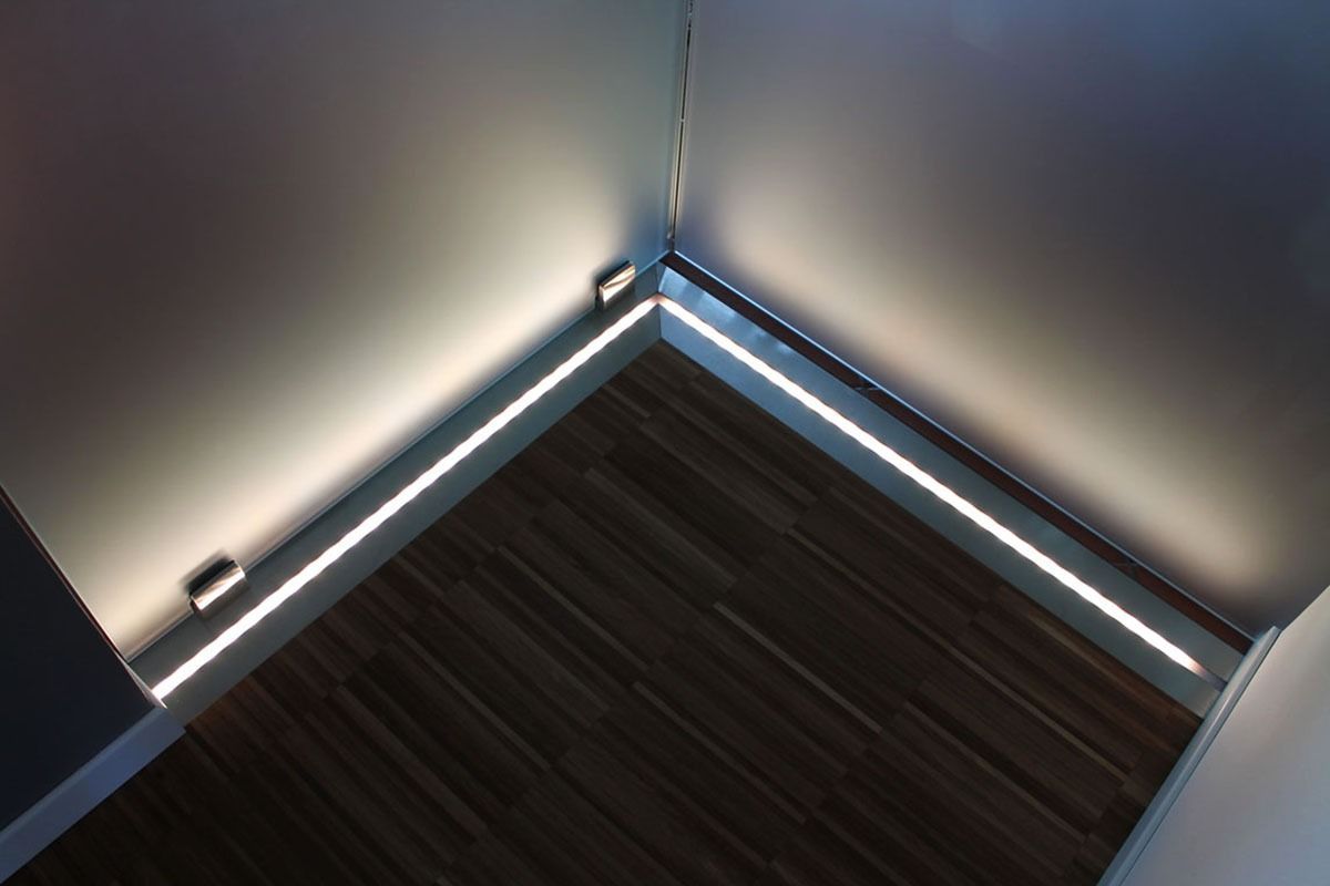 Angolo della stanza con strisce luminose integrate lungo i battiscopa, che illuminano le pareti e il pavimento in legno.