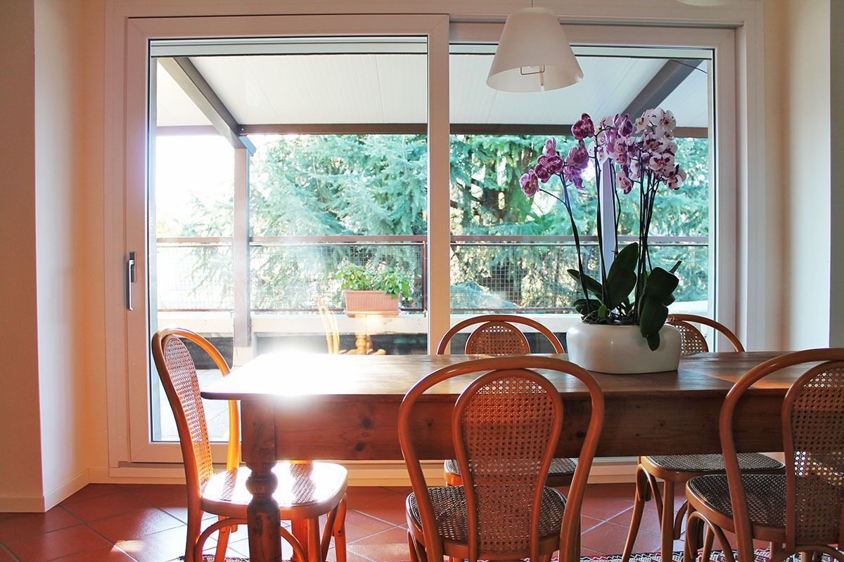 Sala da pranzo con tavolo in legno, sedie in vimini e grande finestra con vista sugli alberi. Orchidea in vaso.