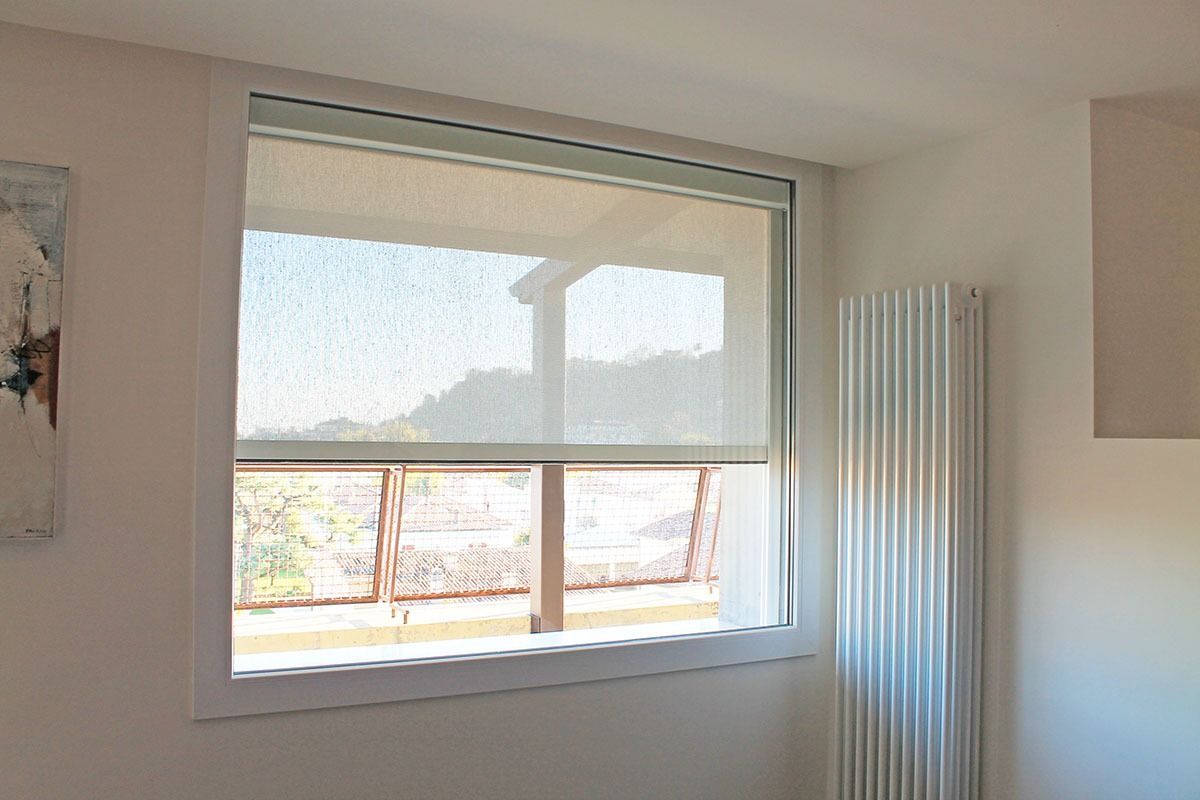Finestra con tenda avvolgibile bianca parzialmente abbassata, che lascia intravedere l'esterno e un termosifone.