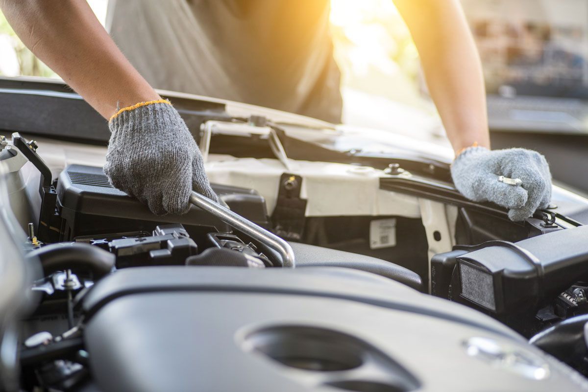 Mechanic in grey gloves working on a car engine outdoors; sunny.