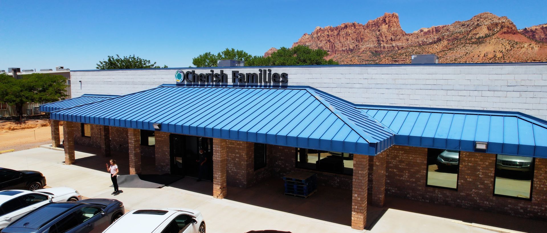 Building with blue roof and "Cherish Families" sign, with cars parked out front, mountains in the background.