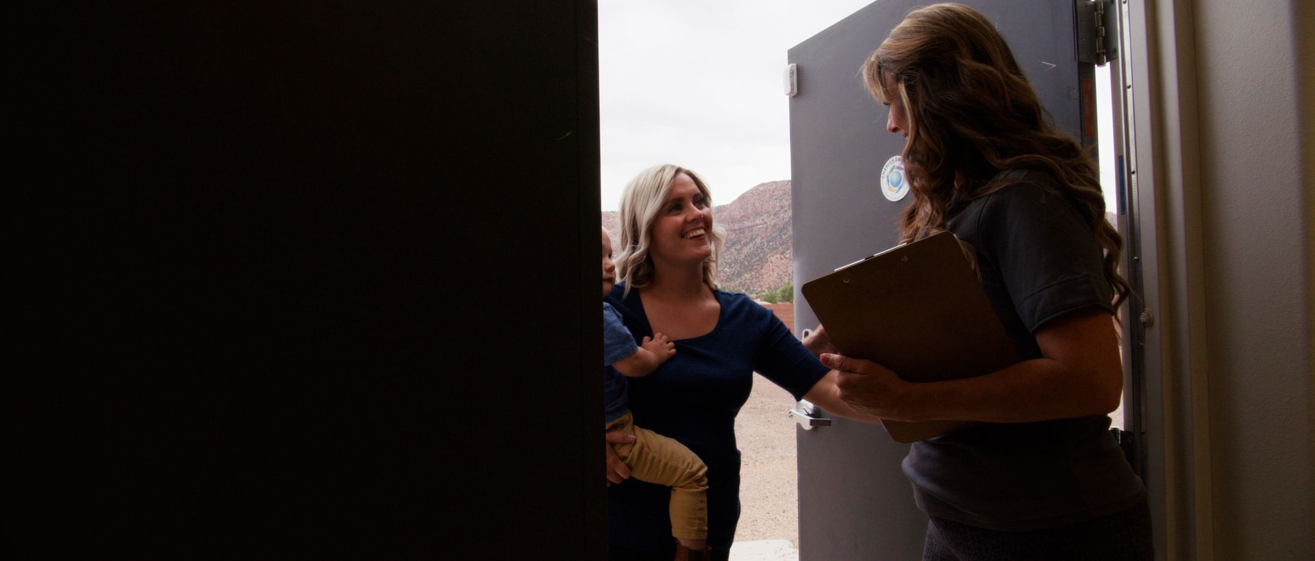 Woman holding a clipboard at a doorway talking to another woman standing outside.