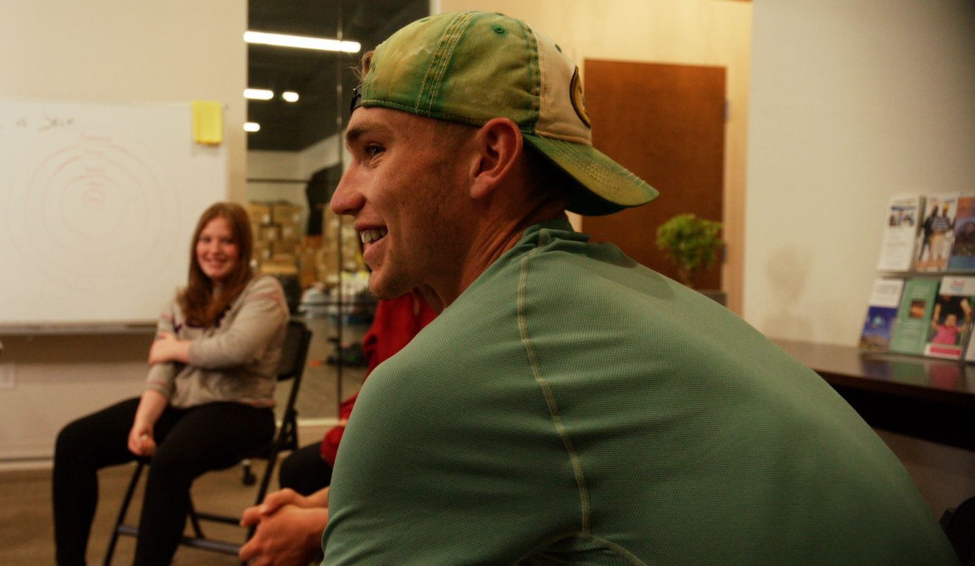 Man in green shirt and hat smiles, seated; woman smiles in background.