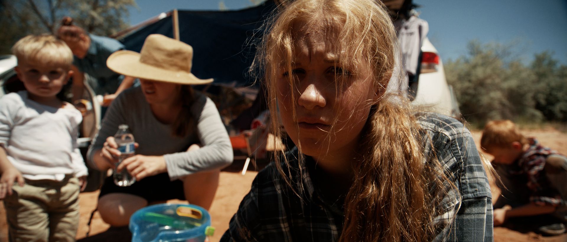 A girl with blonde hair stares forward at a campsite. Family members are in the background, by a tent and car.