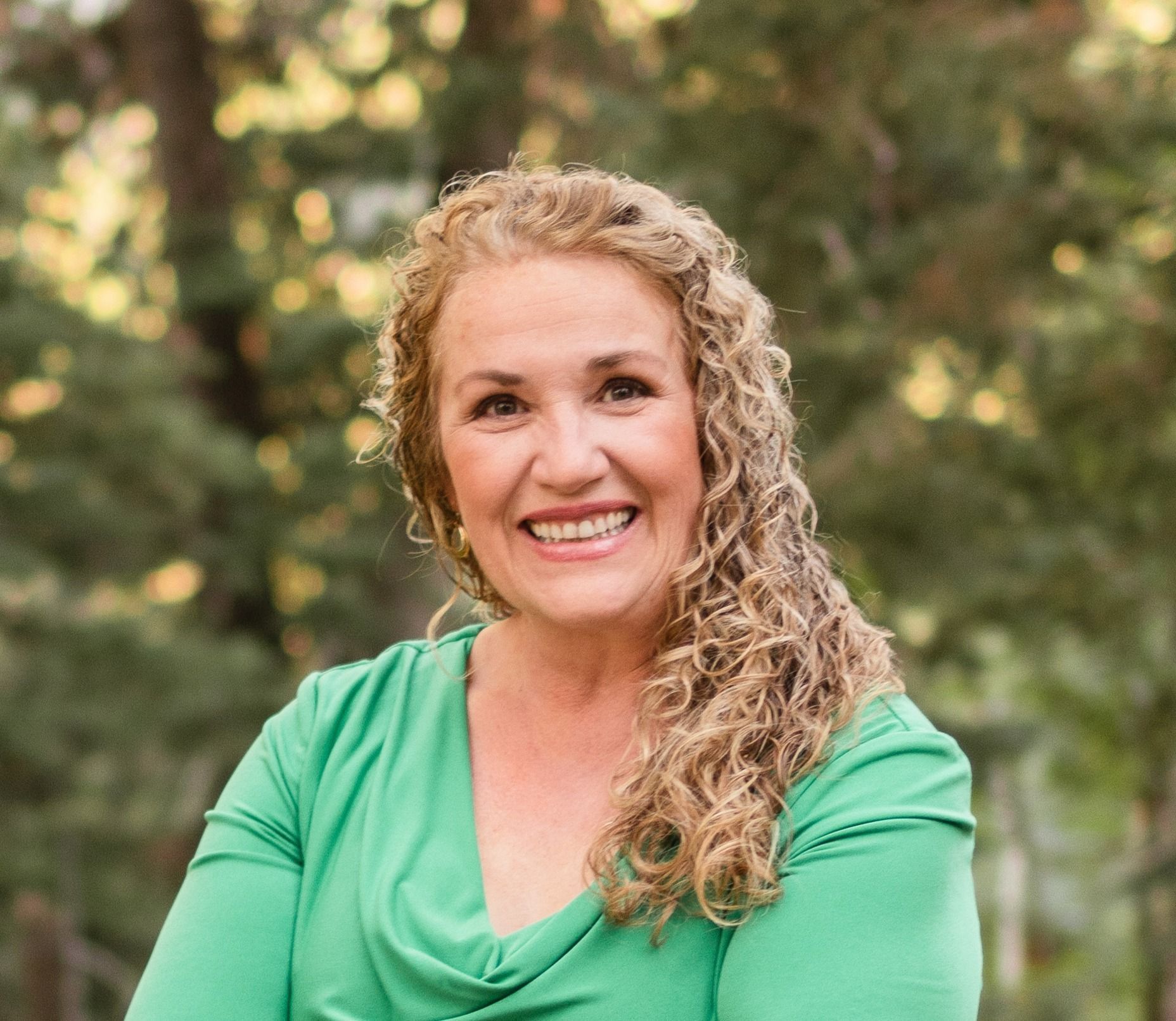 Woman with curly blonde hair, wearing a green top, smiling outdoors.