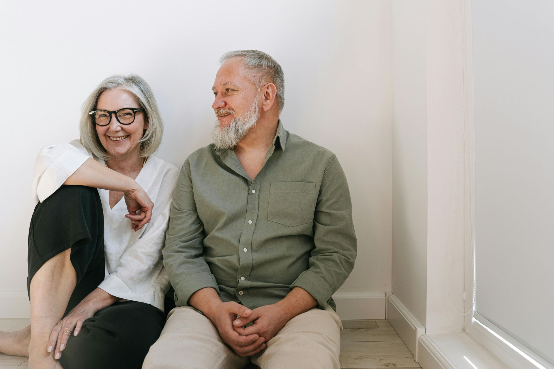 Couple with gray hair smiling, sitting on floor in front of white wall, near window.
