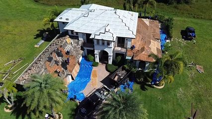 An aerial view of a house under construction with a half-replaced roof, construction debris, and tarps on a green lawn.