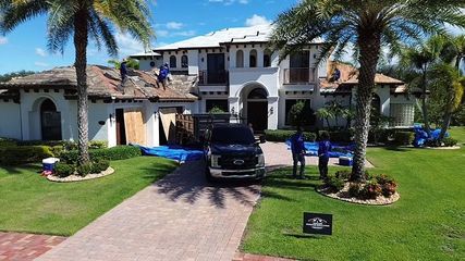 Workers repair the tile roof of a large white house on a sunny day, with a truck parked in the driveway.