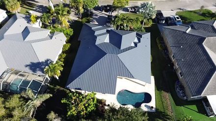 An aerial view shows a house with a dark gray metal roof, a backyard pool, and surrounding neighboring homes and trees.