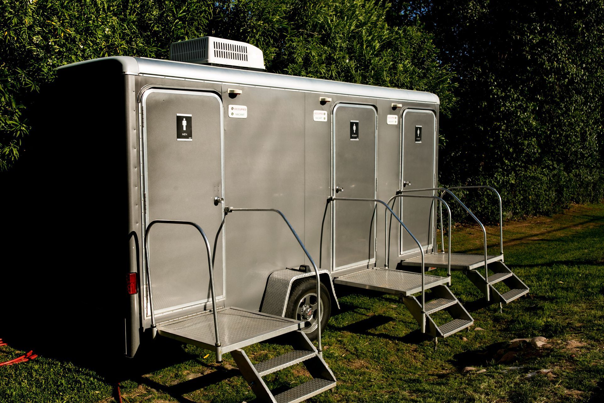Three-stall restroom trailer with steps and safety railings set up outdoors on a grassy area. Three-stall restroom trailer with steps and safety railings set up outdoors on a grassy area.