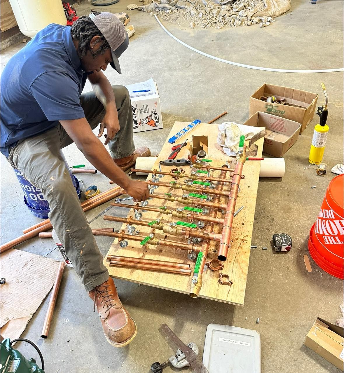 A man is working on a piece of wood in a room with a bucket that says now on it
