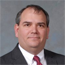 Formal headshot of a man in a suit and red tie against a gray background