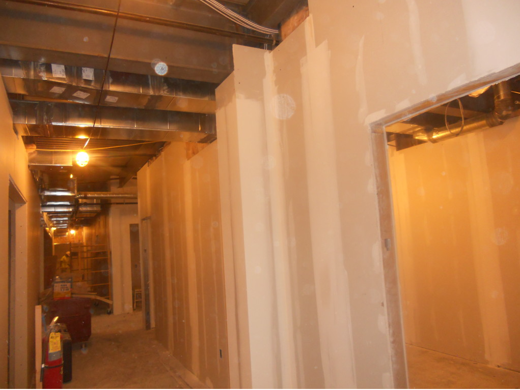 Narrow unfinished hallway with drywall, exposed beams, and hanging lights under construction