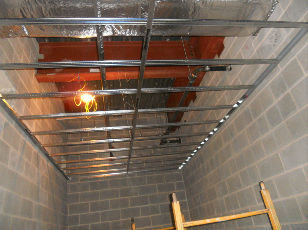 Ceiling under construction with metal framing, orange insulation, and a hanging work light in a cinder block room