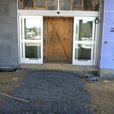 Construction entrance with glass doors, plywood barrier, and gravel path leading up to it