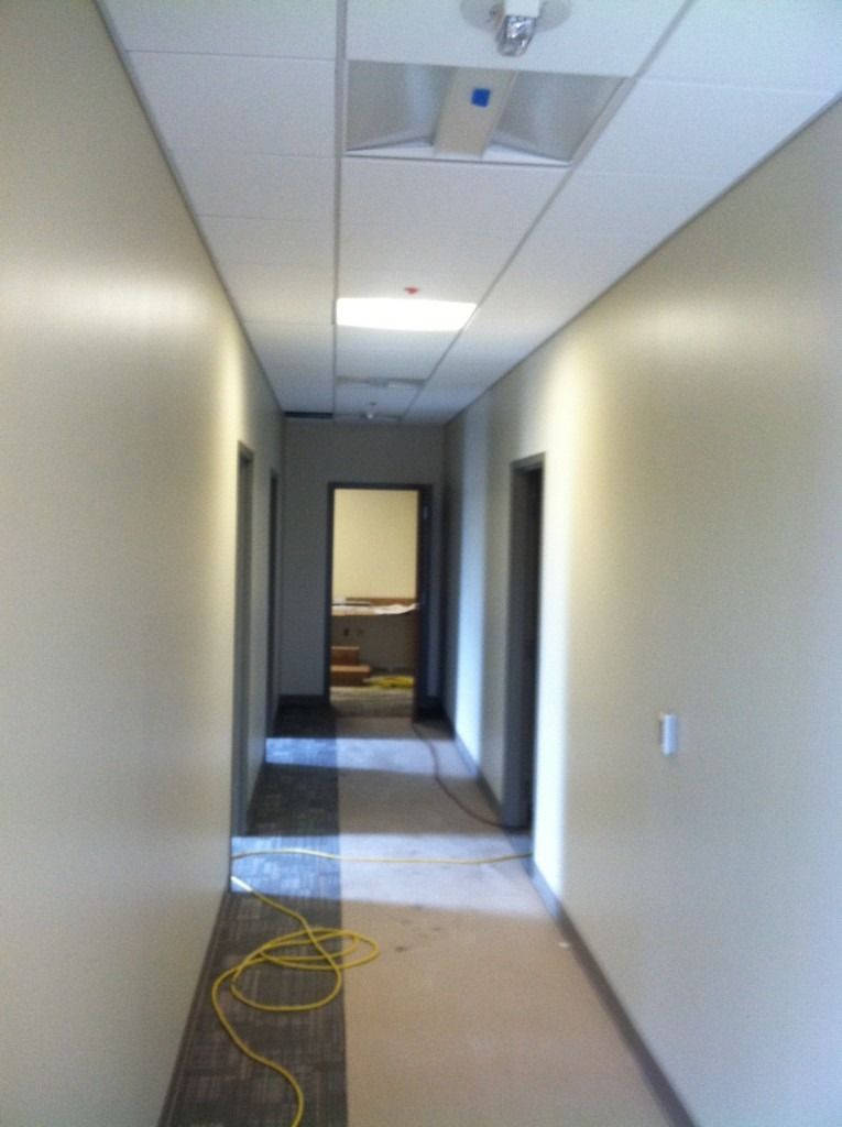 Narrow office hallway with beige walls, ceiling lights, and a yellow cable on the floor near the entrance