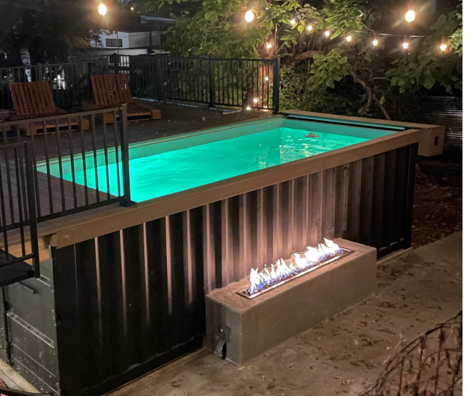 Shipping container pool with fire pit. Dark metal exterior, glowing green water. Black fence and string lights.