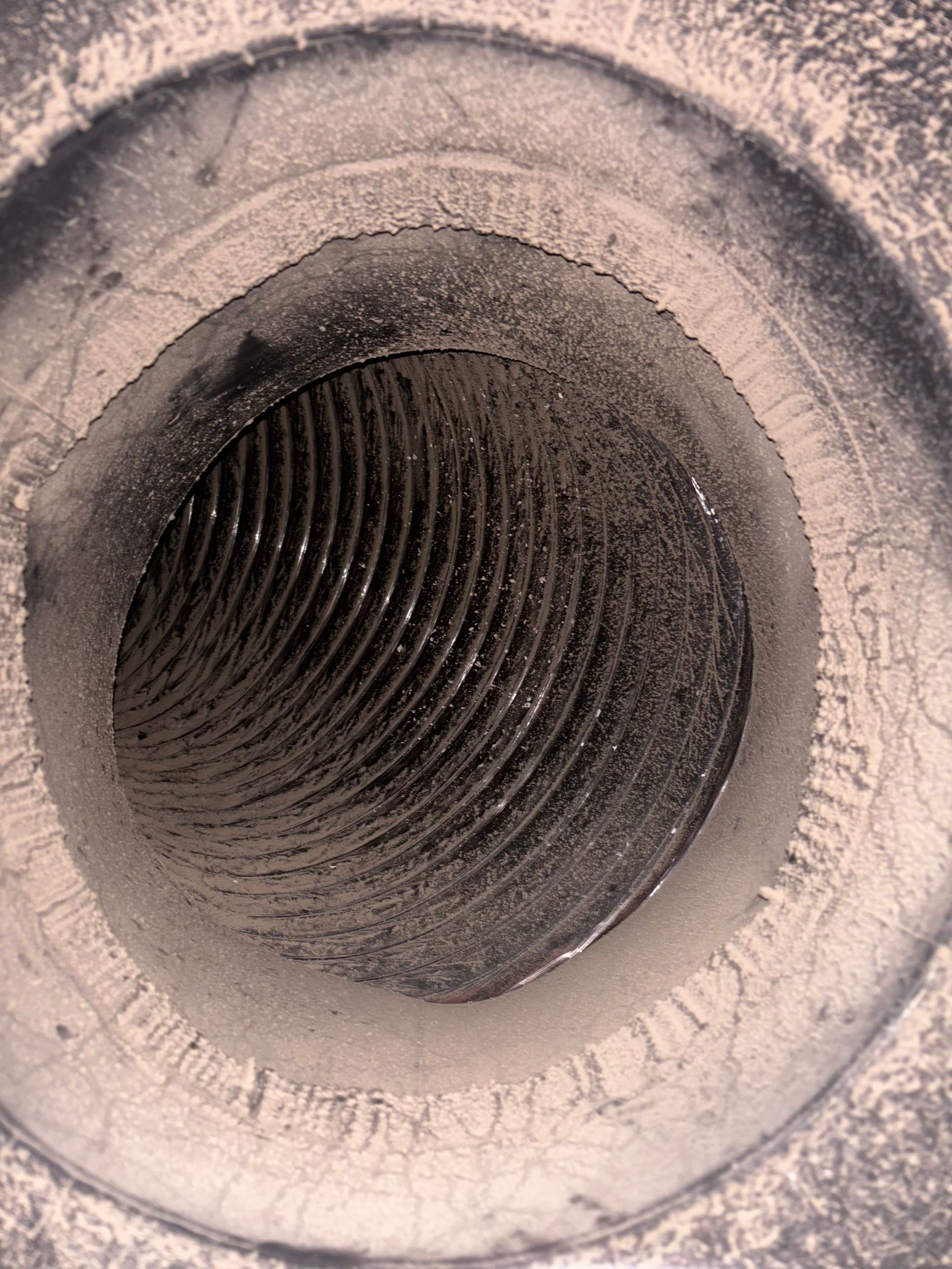 A close-up view down a dark, circular ventilation duct featuring visible ridges and a layer of accumulated gray dust.