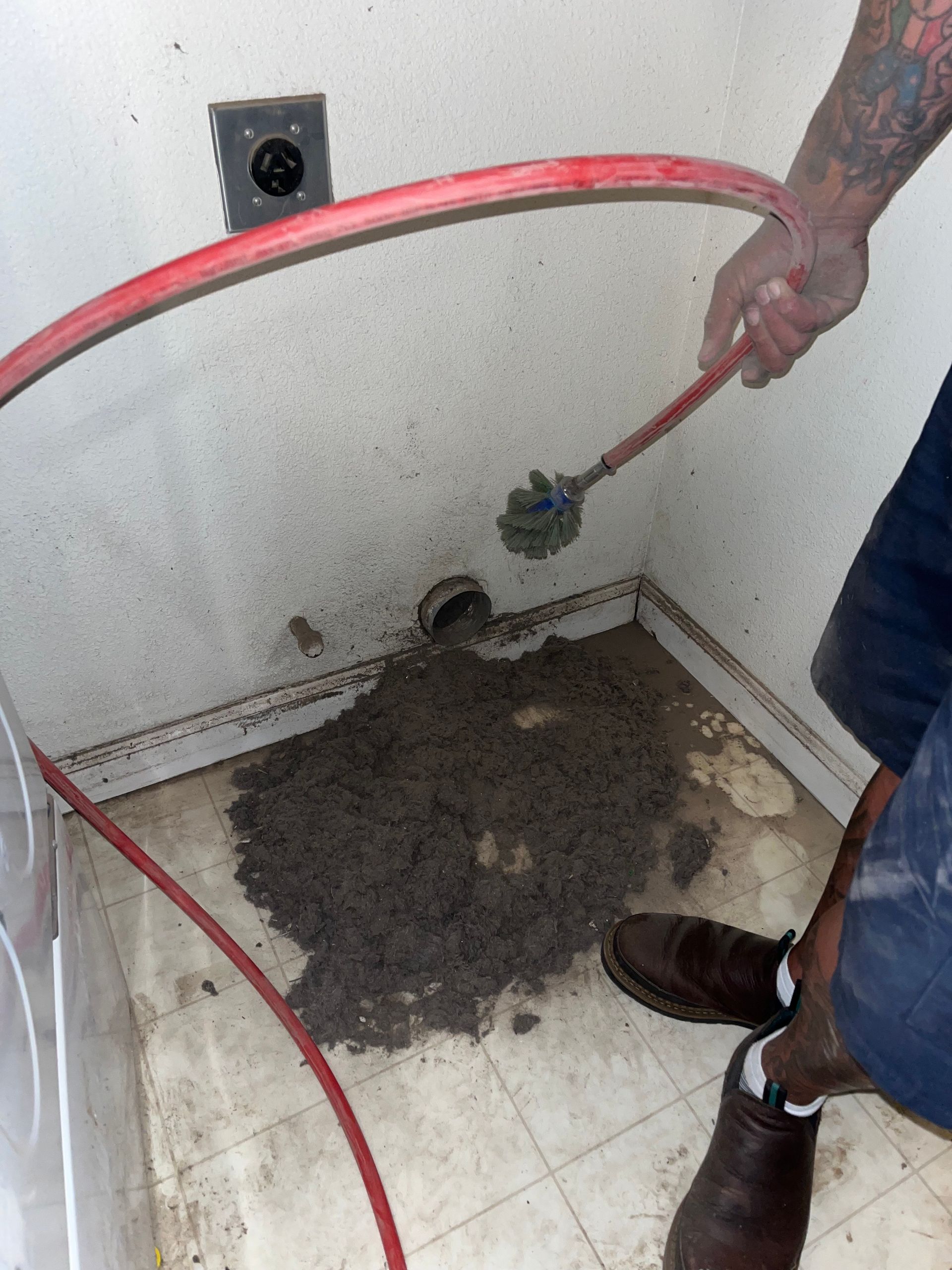 A person uses a long brush attached to a red hose to clean lint from a hole in a wall, with a pile of lint on the floor.