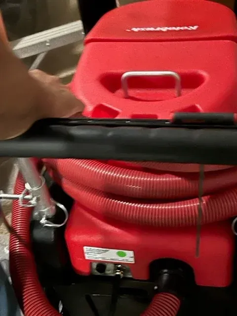 A red commercial carpet cleaning machine with corrugated hoses and a handle, viewed from behind.