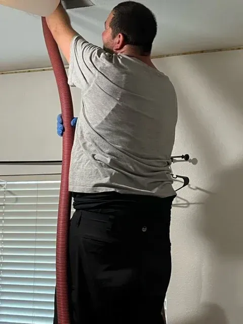 A person standing on a ladder uses a long, red vacuum hose to clean or remove material from a ceiling vent indoors.