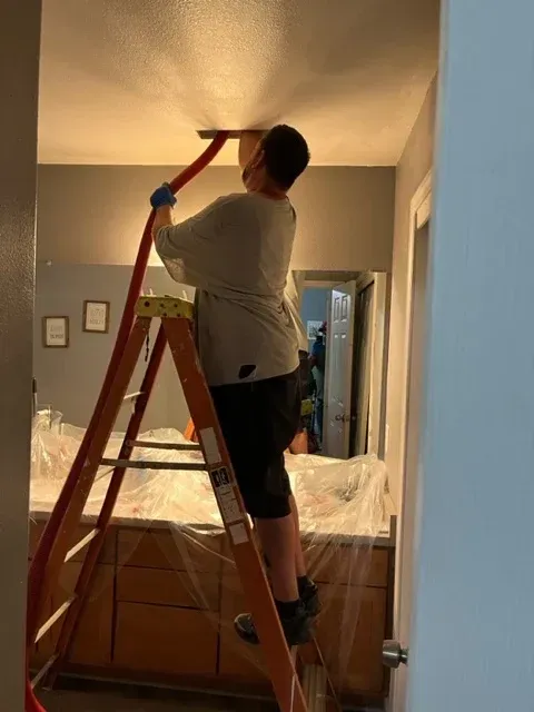 A person on a ladder uses a long-handled tool to sand or scrape a ceiling in a bathroom covered with protective plastic.