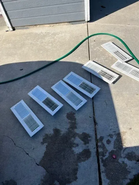 Six white HVAC floor register vents drying on a concrete patio next to a garden hose.