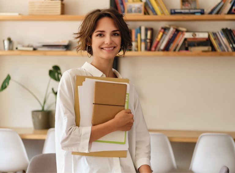A woman in a white shirt is holding a pile of papers and smiling.