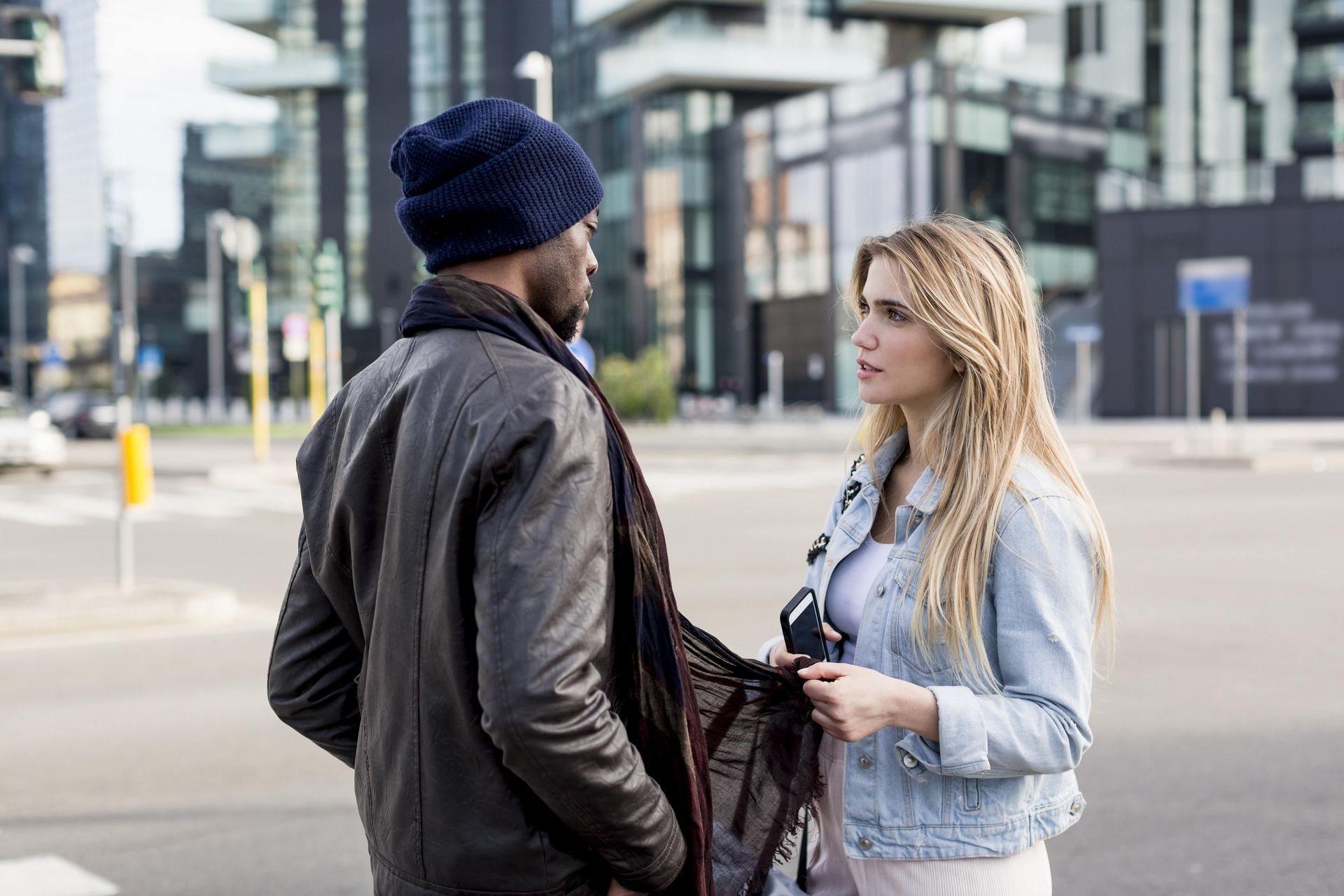 Woman and person wearing a beanie talking on a city street. The woman is holding a scarf. Modern buildings in the background.