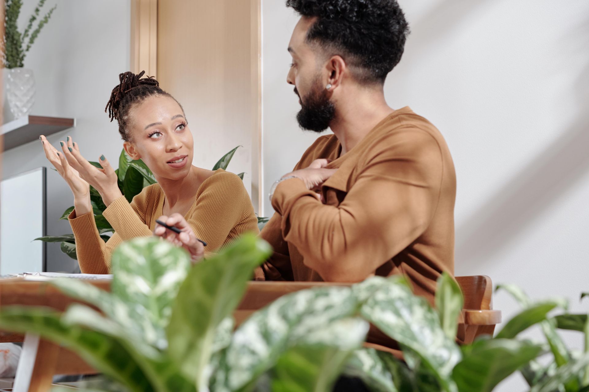 Woman gesturing, man with hand on chest, both looking at each other, discussing at a table.