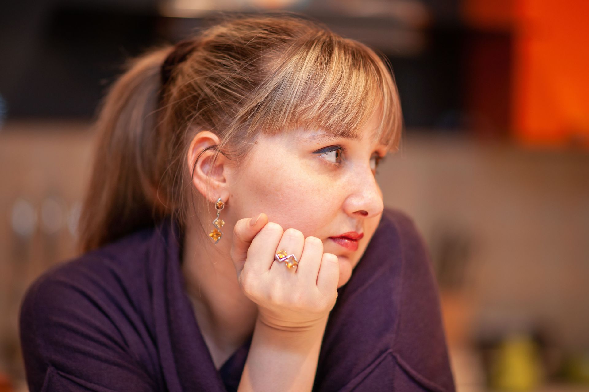 Woman with blonde hair in a ponytail, resting chin on hand, looking off to the side, wearing a purple shirt.