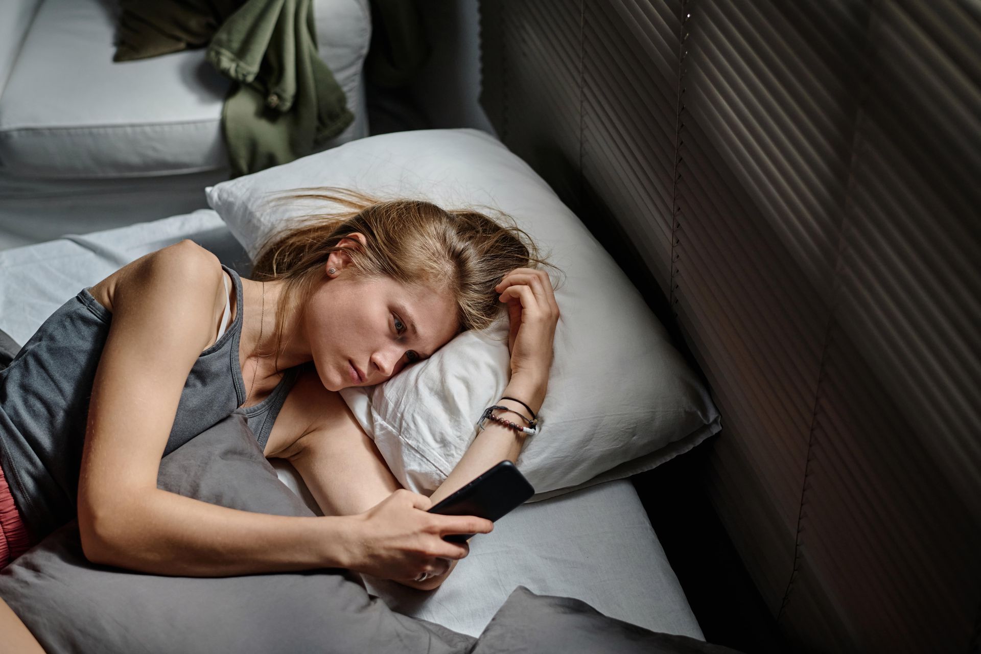 Woman lying in bed, holding phone, looking down with a somber expression.