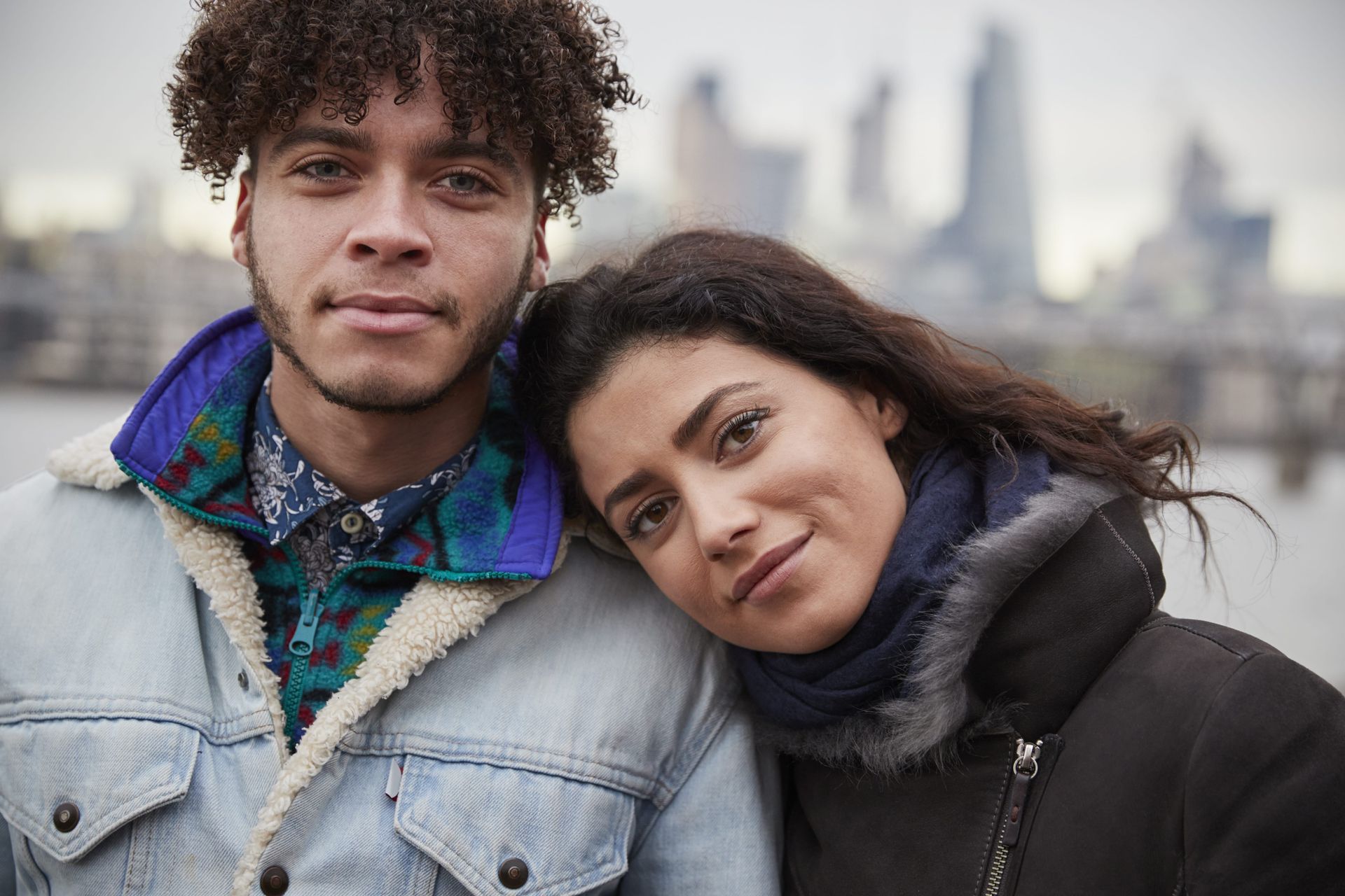 Couple in jackets, smiling; woman's head rests on man's shoulder, London skyline in the background.